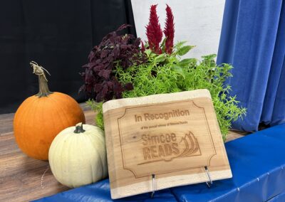 Charcuterie board engraved with "In recognition of the annual winner of the Simcoe Reads with the Simcoe Reads logo below, surrounded by fall decor (pumpkins and foliage).