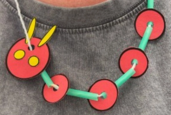A necklace made of green straws and red circular paper pieces, with one piece shaped like a caterpillar’s head featuring yellow antennae and spots.