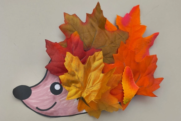 A smiling pink hedgehog cutout with a black nose and eye, its spiky back made of colorful red, orange, yellow, and brown autumn leaves.